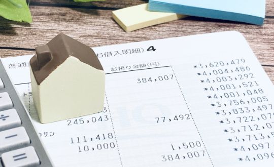 ベージュ色のミニチュアの家が銀行通帳の上に置かれ、その左側に電卓が見える様子